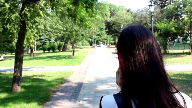 有魅力的女孩穿着白衬衫，在公园里散步时打电话。年轻漂亮的女孩一边在公园里散步一边打电话。视频素材