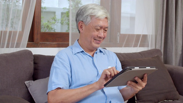 亚洲老年男性在家里使用平板电脑。亚洲资深华人男性躺在家里客厅的沙发上上网搜索关于如何保持健康的信息。视频素材