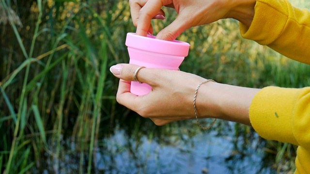 女性双手打开一个装有粉色硅胶月经杯的容器，把它展示出来，并把里面放在绿色的自然背景和湖泊上。妇女健康理念，零废物替代品视频素材