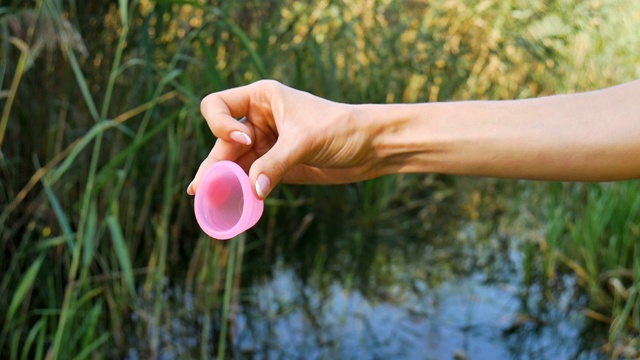 粉色硅胶月经杯在女性的手中，绿色的自然背景与湖泊。妇女健康理念，零废物替代品视频素材