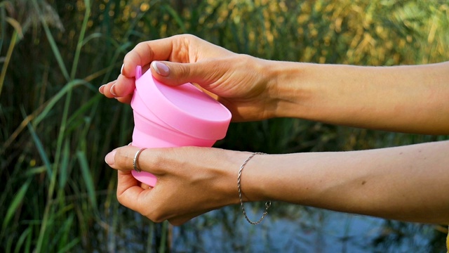 女性的手正在展示如何使用一个粉红色的硅胶月经杯站在绿色的自然背景和湖泊。妇女健康理念，零废物替代品视频素材