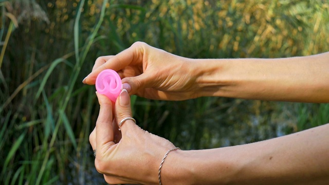 女人拿着一个粉红色的月经杯站在绿色的自然背景与湖泊。女性卫生的概念视频素材
