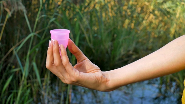 特写女性手与粉红色硅胶月经杯站在绿色自然背景与湖泊。零废物替代月经周期视频素材