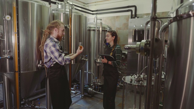 啤酒厂的工人看着玻璃管里新酿的啤酒并讨论着它。男性和女性啤酒酿造者在啤酒厂测试啤酒。4 k。中小企业的概念视频下载