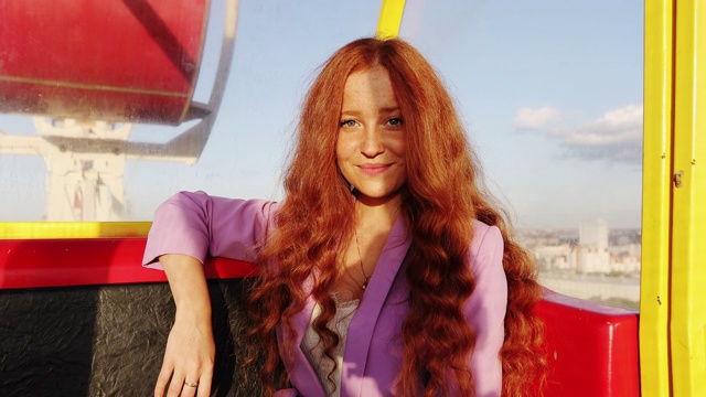 太阳亲吻女孩，欣赏夕阳。一位有着红色长发的年轻女子正坐在缆车或摩天轮舱内。视频下载