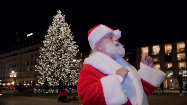 圣诞老人站在广场上，圣诞树照亮了夜晚视频下载