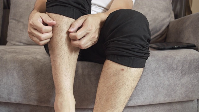 4k分辨率亚洲男子坐在家里的沙发上用手抓挠他痒的皮肤腿。蚊虫叮咬后疼痛。医疗保健理念。视频素材