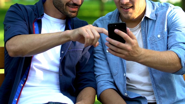 两个朋友坐在公园的长椅上用智能手机看有趣的视频，特写视频素材