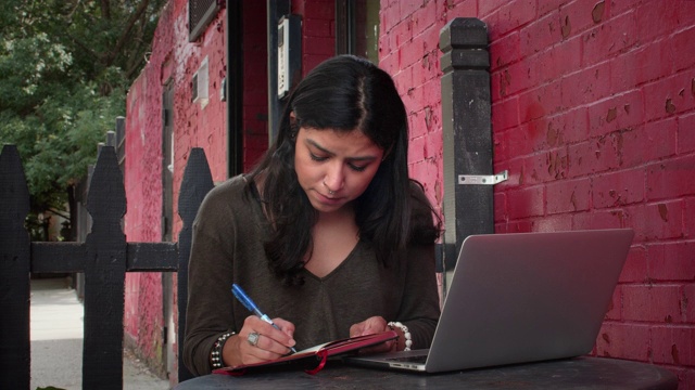 女性千禧一代在笔记本电脑上工作，在户外的预约簿上书写café视频下载