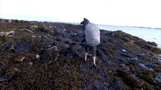 两个人扛着设备越过多岩石的海岸。视频下载