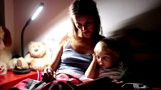妈妈晚上在家给蹒跚学步的儿子读书，床头灯开了，妈妈和宝宝拥抱在一起视频素材
