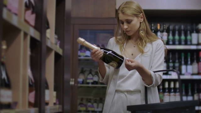 女孩在商店里看酒瓶上的标签买酒，然后在酒架上选择了一种超市里的酒精饮料视频素材