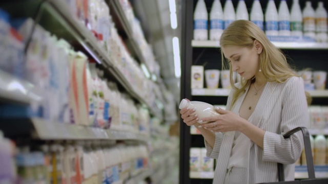 一个女人从超市冰箱里拿出一瓶牛奶，看了看标签和产品成分。把牛奶放在篮子里视频素材