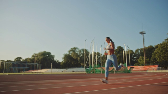 美丽的健身女人在浅蓝色运动上衣和打底裤跑得非常快在一个户外体育场。在一个温暖的夏日午后，她在短跑。运动员在跑道上进行运动练习视频下载