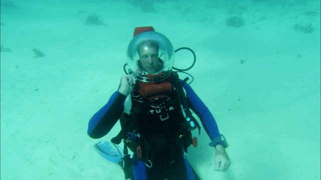 一名戴水肺的潜水员坐在沙质海床上。视频下载