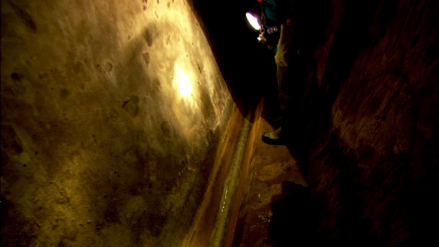 一名下水道工人拿着手电筒走过一条隧道，停在墙上的一个洞口前。视频下载