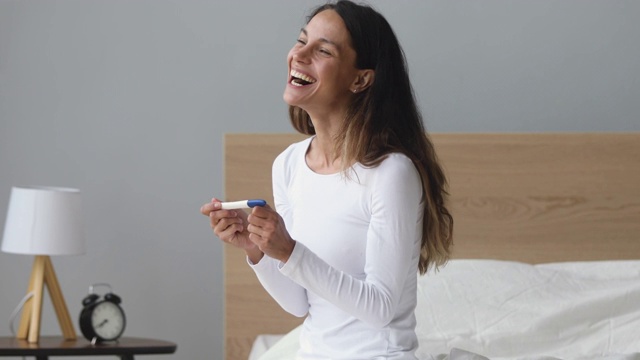 兴奋的年轻女子坐在床上抱着阳性妊娠测试视频素材