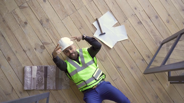 一个戴着建筑工人头盔，穿着绿色夹克的快乐微笑的男人躺在地板上，用胜利的手势向上看视频素材
