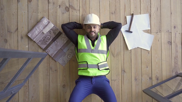 快乐的建设者在头盔和绿色夹克躺在地板上向上看。锤子和画就在他旁边。视频素材