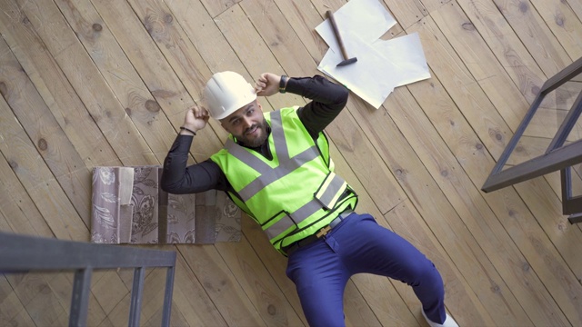 快乐微笑的建设者在头盔和绿色夹克躺在地板上显示胜利的手势和向上看。视频素材