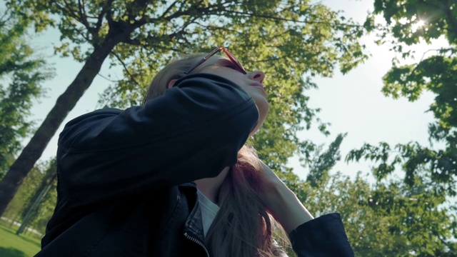 美丽的年轻女孩模特与长头发在公园的相机摆姿势，在绿色的树的背景。寻找一个好的自拍姿势。穿着皮夹克视频下载