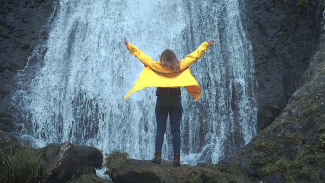 穿着黄色雨衣的年轻女孩徒步旅行者走向高地上的瀑布，举起双手，享受自然和生活。在山里旅行，在旅行中冒险。生活方式的概念视频素材