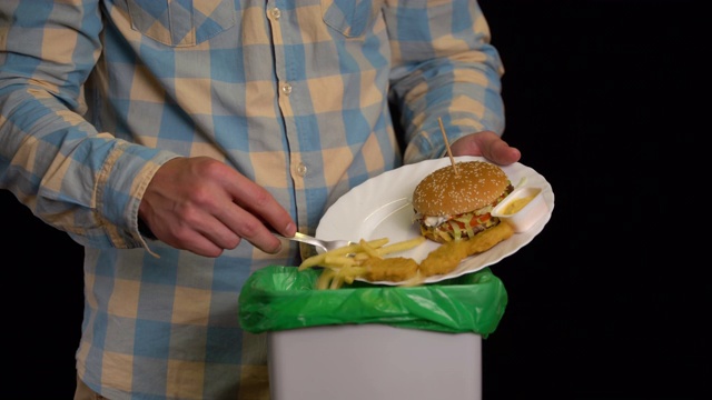 一个男人用盘子刮一个汉堡，炸薯条，鸡块，奶酪酱到垃圾桶里。视频素材
