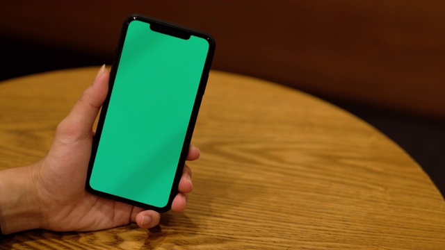 一只手拿着绿色屏幕的智能手机放在有图案的木桌上。视频素材