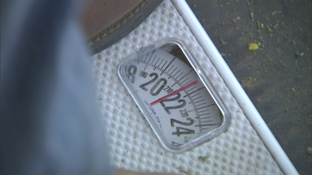 一个男人站在秤上，显示的是220磅。视频下载