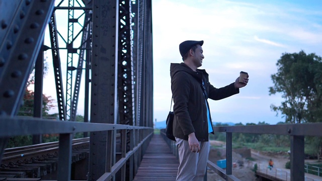 亚洲游客穿着大衣，喝着外卖咖啡杯，站在黑色铁桥铁路背景下。视频下载