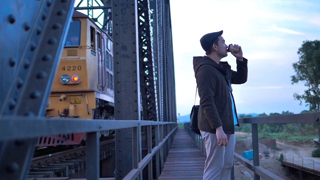 穿着大衣的亚洲游客在黑色铁桥铁路背景下喝着外卖咖啡杯。视频下载