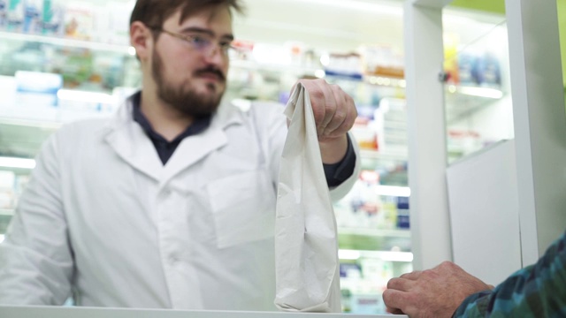 留着胡须的年轻药剂师把装有药物的白色包裹递给顾客。视频下载