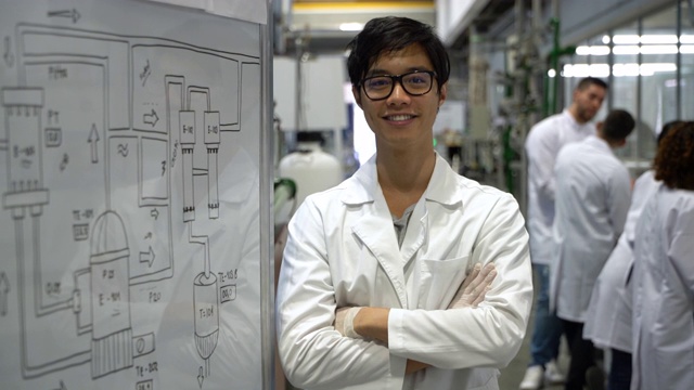 一个快乐的亚洲学生站在实验室的白板旁边，对着镜头微笑，双臂交叉视频下载