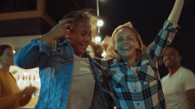 一群朋友在街头美食汉堡店外举行聚会。两个美丽的女孩随着音乐拥抱跳舞。这是现代街区的夜晚。每个人都是幸福的，充满喜悦。视频素材