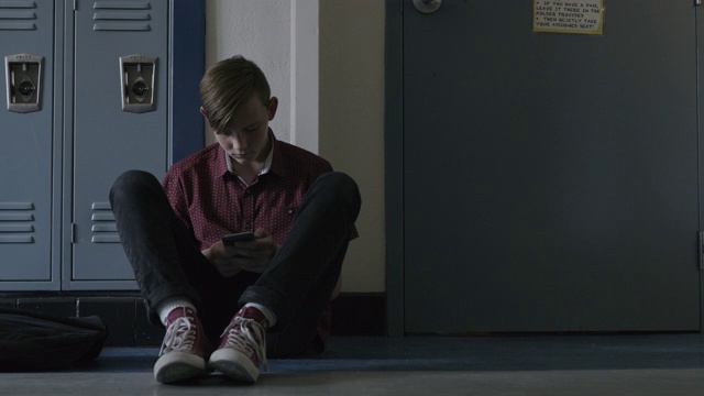 美国犹他州普罗沃，一个孤独的男孩坐在学校走廊的地板上使用手机视频下载