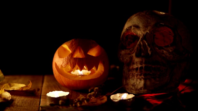 小长着牙齿的南瓜杰克和一个在烛光下发光的眼睛的人的头骨放在一张木桌上视频下载