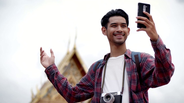 年轻英俊的亚洲男子独自站在街上用智能手机与朋友视频聊天，背景是泰国的寺庙。快乐享受休闲旅游生活方式。视频下载