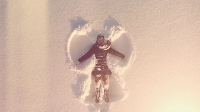 空中俯瞰年轻快乐的女人躺在雪地里，双手和双腿在阳光下做雪天使视频下载
