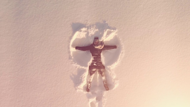 空中俯瞰年轻快乐的女人躺在雪地里，双手和双腿在阳光下做雪天使视频素材