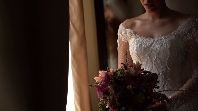 新娘手持花束，在婚礼前透过窗户看视频素材
