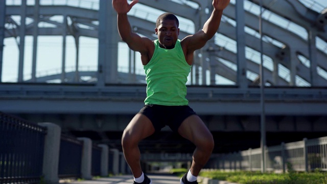 多利拍摄的决心良好肌肉的非洲年轻运动员在城市桥梁上进行蛙蹲跳练习视频下载