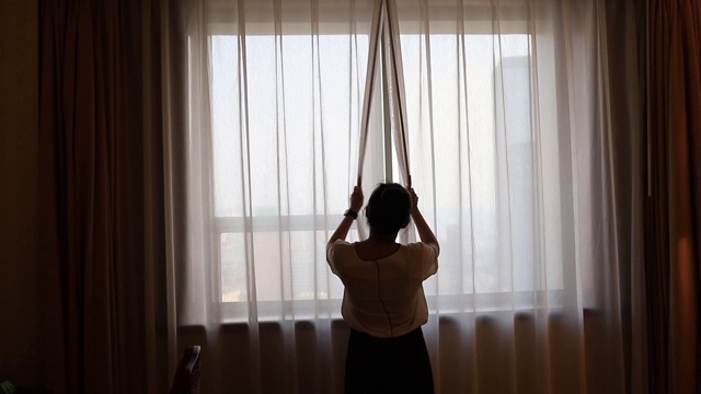 亚洲女性住在酒店房间里。打开房间里的窗帘，看外面的景色。假日旅游概念。视频下载