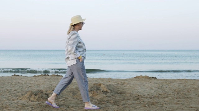 清晨日出时，戴着草帽的快乐老妇人走在海滩上。视频素材