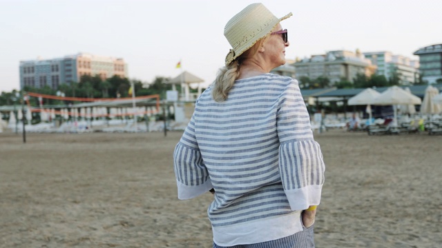 清晨日出时分，一位戴着草帽的快乐的老妇人走在海滩上。视频素材