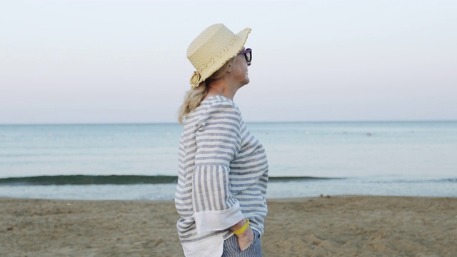 清晨日出时，戴着草帽的快乐老妇人走在海滩上。视频素材