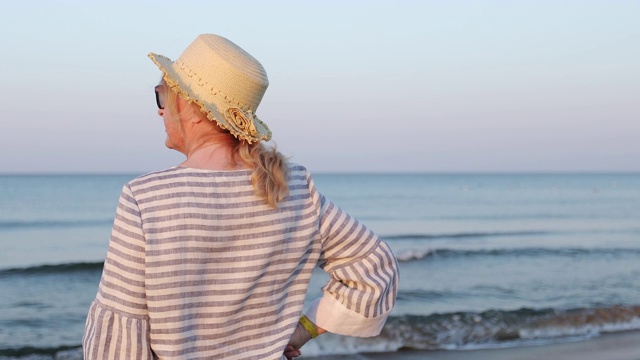 清晨，一位戴着草帽的老妇人站在海滩上。视频素材