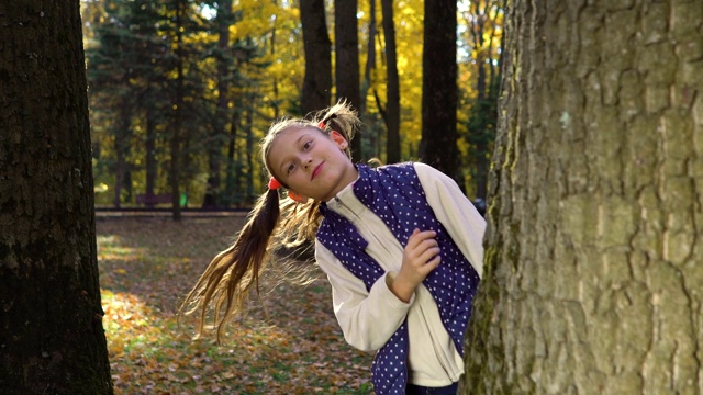 可爱的女孩在秋天公园的树后面玩捉迷藏的慢镜头视频素材