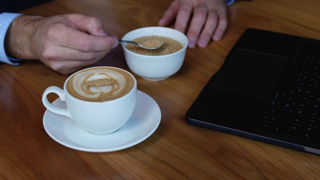 男子在一杯咖啡中搅拌糖。在桌上一台笔记本电脑旁边视频素材