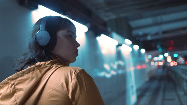在日本东京，一名年轻女子戴着耳机和兜帽夹克在夜间的隧道中行走视频下载