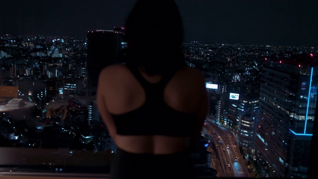 在日本东京，年轻女子穿着运动胸罩戴着耳机，看着窗外的夜晚视频下载